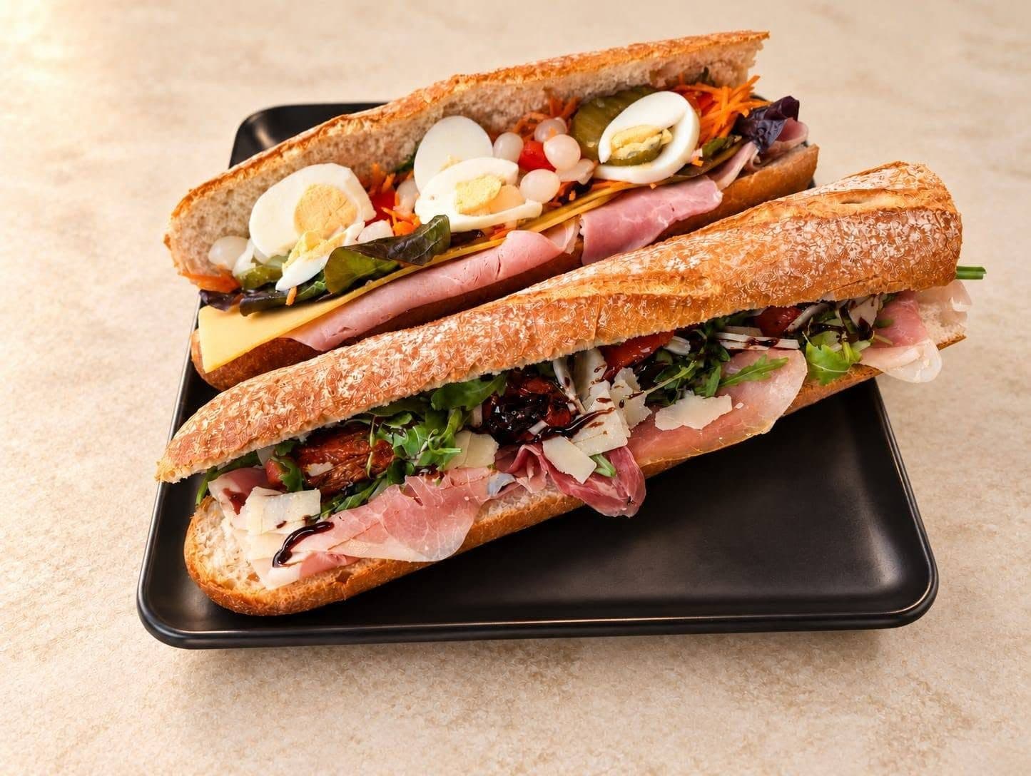 Deux baguettes garnies posées sur un plateau noir, sur le comptoir de la sandwicherie